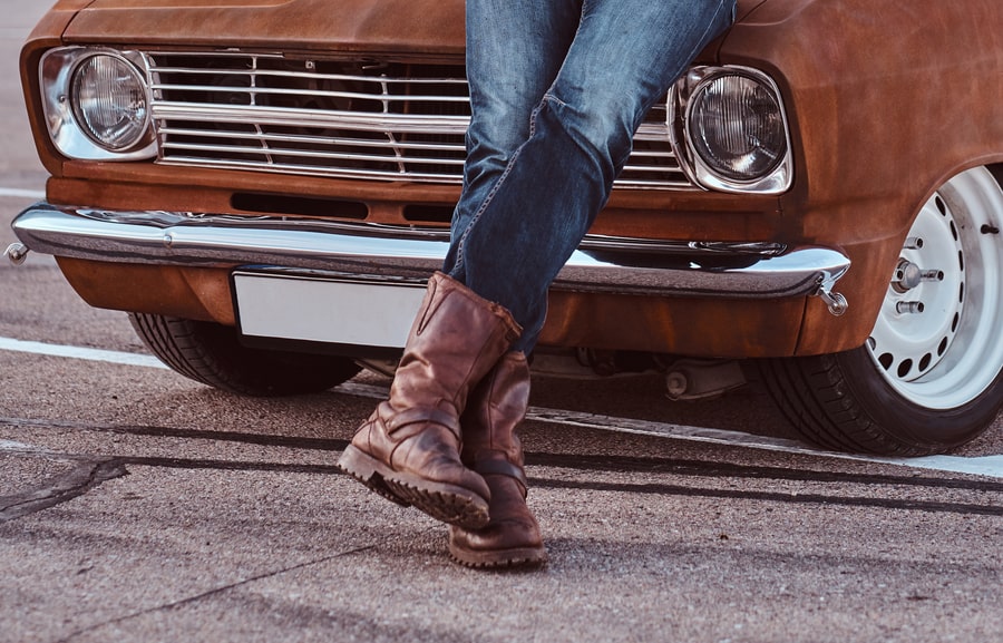 Male in jeans and boots leaning on retro car in the city parking.